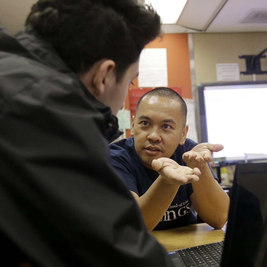 In this April 19, 2016, photo, high school teacher Dennis Caindec, right, talks with a student during a twelfth-grade senior seminar class at San Francisco International High School in San Francisco. While some districts in numerous states have discouraged migrant minors from Central America from enrolling in their schools, San Francisco International High School accommodated its youths by rewriting young-adult novels at a basic level to spark the newcomers' interest in reading. (AP Photo/Jeff Chiu)