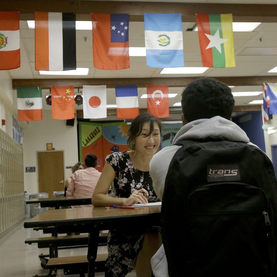 In this April 19, 2016, photo, high school teacher Tara Hobson talks with a student in the school cafeteria at San Francisco International High School in San Francisco. Some districts have gone to extraordinary lengths to accommodate migrant students, who often come to join relatives, sometimes escaping criminal gangs or extreme poverty. San Francisco International High School rewrote young-adult novels at a basic level to spark the newcomers' interest in reading. (AP Photo/Jeff Chiu)